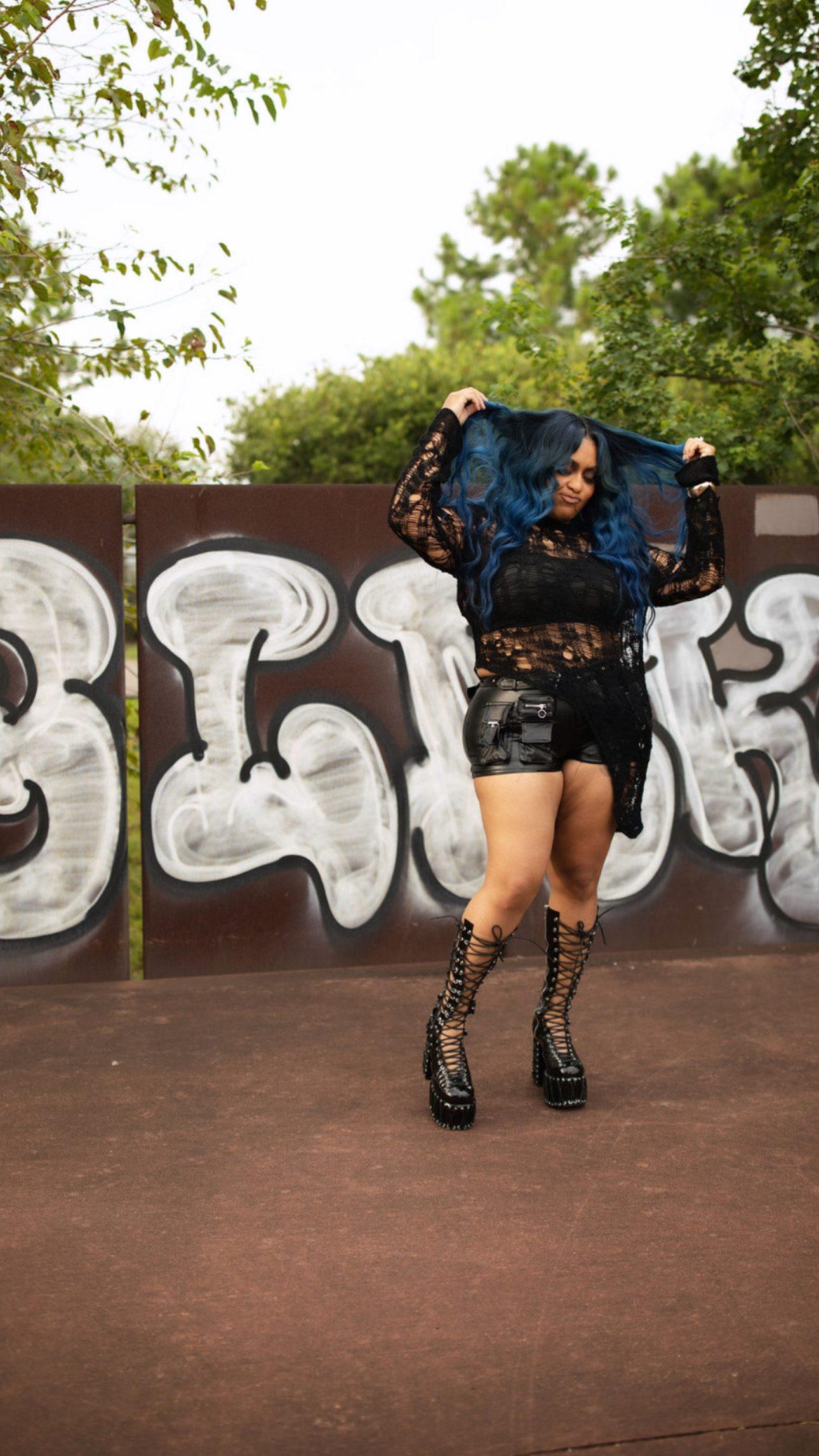 Jess lifting both hands into a custom blue wavy unit, showing its length and movement, in front of a white graffiti wall