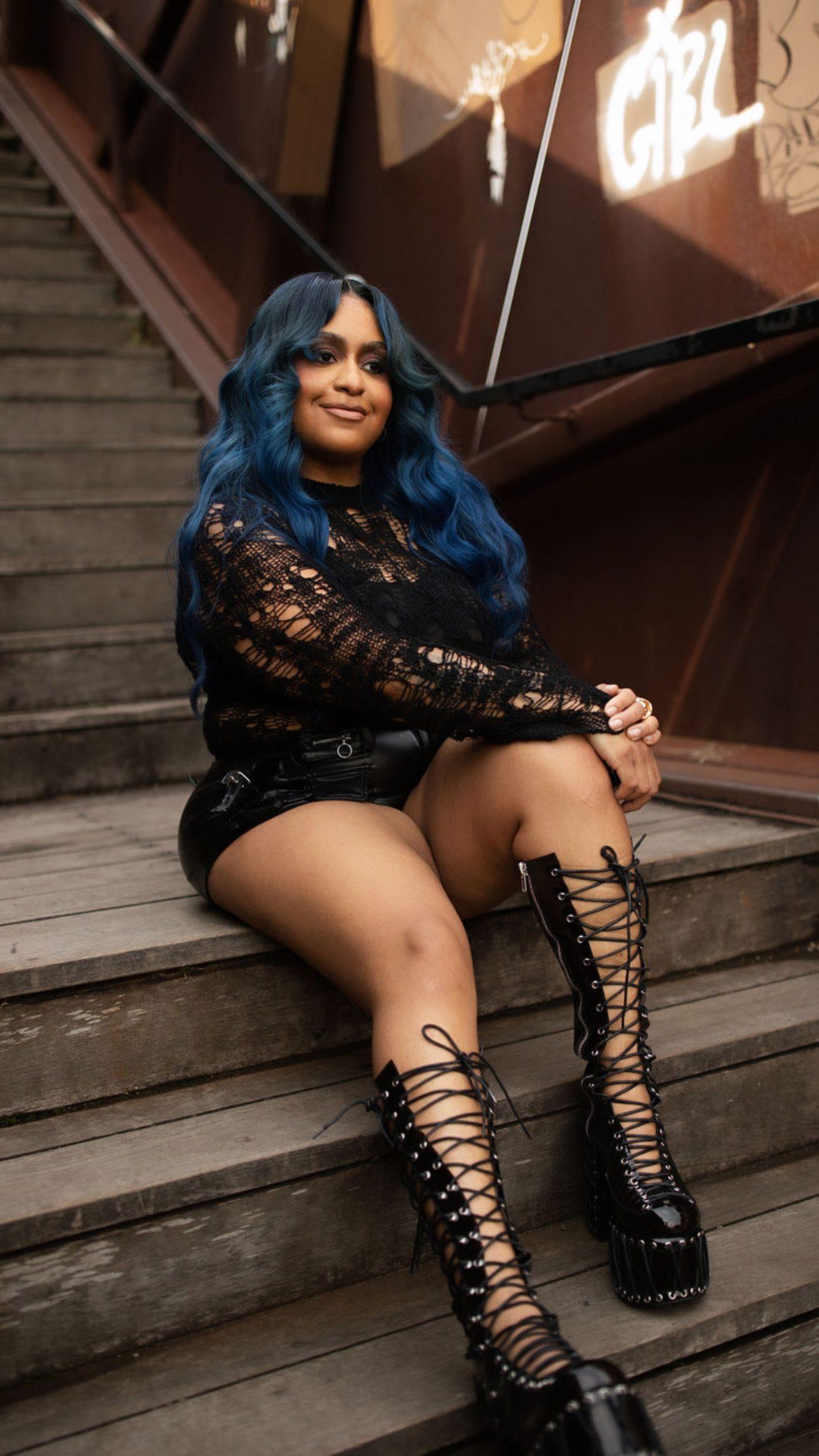 Jess seated on open metal stairs in a custom blue wavy unit and lace-up platform boots, relaxed and smiling